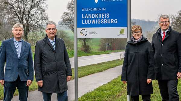 Die Aufnahme zeigt vier Personen neben einem Willkommensschild, das "Willkommen im Landkreis Ludwigsburg" anzeigt. Im Hintergrund sind eine Straße und Bäume zu sehen. Die Personen tragen formelle Kleidung und stehen auf einer Wiese.