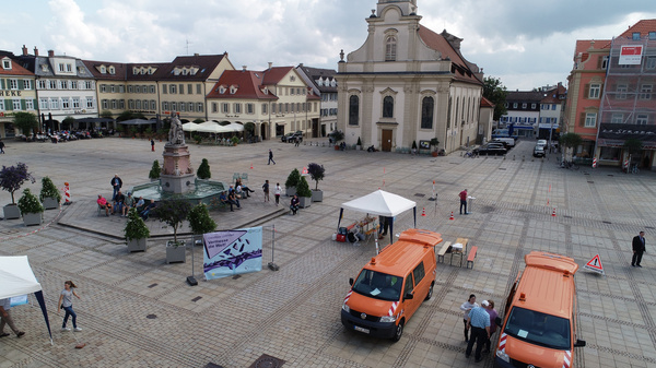 A public square features a fountain surrounded by small trees, with a church in the background. Several people are walking, and two orange vans are parked nearby. Tents are set up, indicating an event or gathering in the area. The atmosphere appears lively and communal.