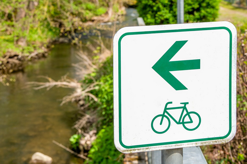 Ein Verkehrszeichen zeigt einen grünen Pfeil nach links und ein Fahrradsymbol an. Im Hintergrund fließt ein kleiner Bach, umgeben von Bäumen und Sträuchern. (Text KI generiert)