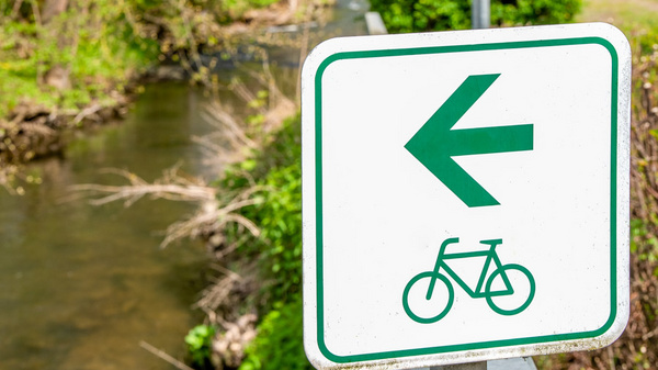 Ein Verkehrszeichen zeigt einen grünen Pfeil nach links und ein Fahrradsymbol an. Im Hintergrund fließt ein kleiner Bach, umgeben von Bäumen und Sträuchern. (Text KI generiert)