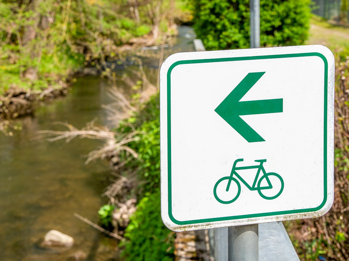 Ein Verkehrszeichen zeigt einen grünen Pfeil nach links und ein Fahrradsymbol an. Im Hintergrund fließt ein kleiner Bach, umgeben von Bäumen und Sträuchern. (Text KI generiert)