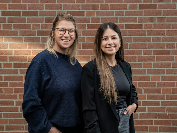 Zwei Frauen stehen lächelnd vor einer Backsteinwand. Die Frau links trägt ein dunkles Sweatshirt und hat brünette, leicht gewellte Haare. Die Frau rechts trägt ein schwarzes Oberteil und hat langes, glattes Haar. Beide wirken freundlich und entspannt.