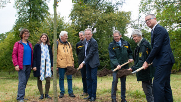 A group of eight individuals stands around a freshly dug hole in a grassy area, engaged in planting a young tree. They are dressed in casual outdoor clothing, smiling and holding shovels. Trees and greenery are visible in the background, suggesting an environmental or community initiative.