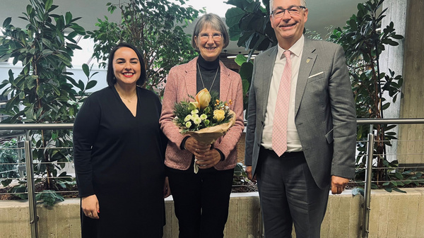 Drei Personen stehen in einem Innenraum mit Pflanzen im Hintergrund. Eine Frau in der Mitte hält einen Blumenstrauß und lächelt, während die anderen beiden Personen neben ihr stehen und ebenfalls lächeln.