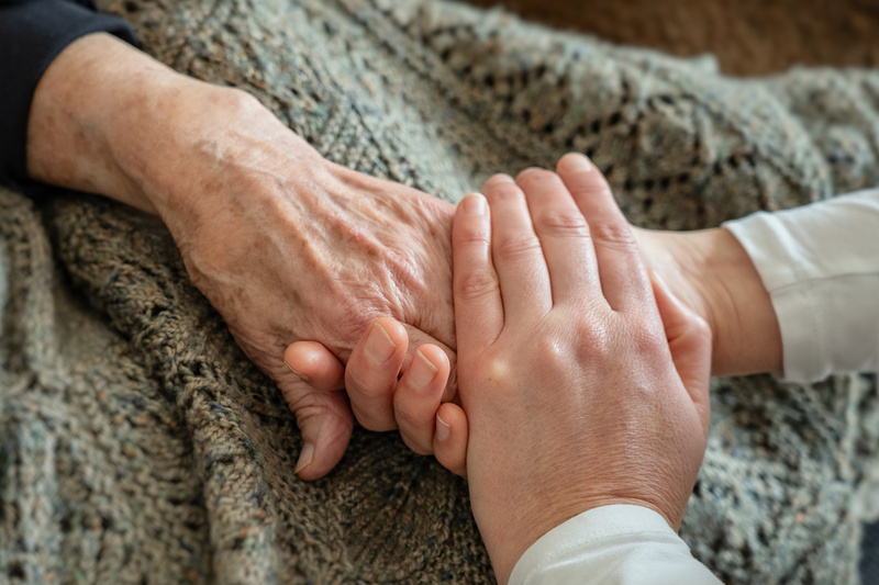 Zwei Hände halten sich liebevoll. Die eine Hand ist älter mit ausgeprägten Adern, während die andere jünger und zarter ist. Im Hintergrund ist eine Decke zu sehen. Die Geste vermittelt Wärme und Zuneigung zwischen den Generationen.