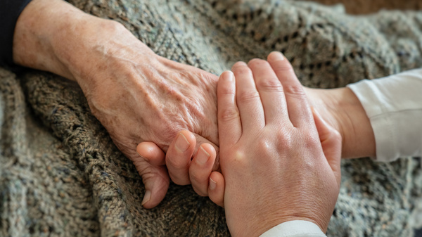 Two hands are gently clasped together, one older and wrinkled, the other younger and smoother. They rest on a textured blanket, symbolizing connection, care, and support between generations.