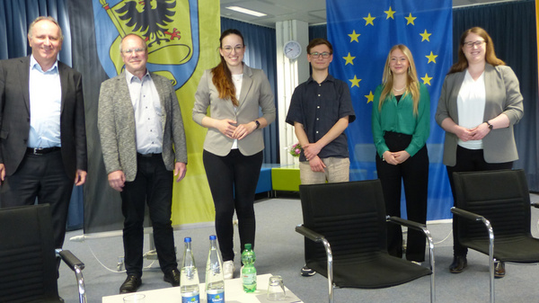 Immagine che mostra un gruppo di sei persone in piedi accanto a bandiere europee e tedesche. Sono sorridenti e vestiti in modo formale. In primo piano ci sono tavoli con bottiglie d'acqua e microfoni. L'ambiente è un ufficio moderno, con tende e pareti colorate.