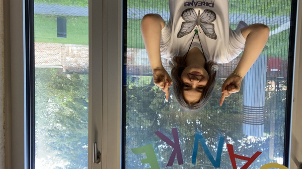 Eine lächelnde Person zeigt mit beiden Händen nach unten auf das Wort "DANKE", das farbenfroh am Fenster geschrieben ist. Im Hintergrund sind Bäume und Rasen sichtbar.