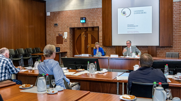 Eine Sitzung findet im Landratsamt statt. Zwei Redner sitzen an einem Tisch vor einer Präsentation, während mehrere Zuhörer in einem Konferenzraum aufmerksam zuhören. Auf den Tischen stehen Getränke und Snacks.