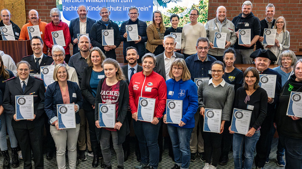 Eine Gruppe von Menschen steht lächelnd zusammen in einem Raum. Sie halten Urkunden in den Händen und stehen vor einem Banner, das "Herzlich Willkommen zum Fachkongress Kinderschutzverein" zeigt. Die Anordnung ist freundlich und feierlich, was den Anlass des Treffens unterstreicht.