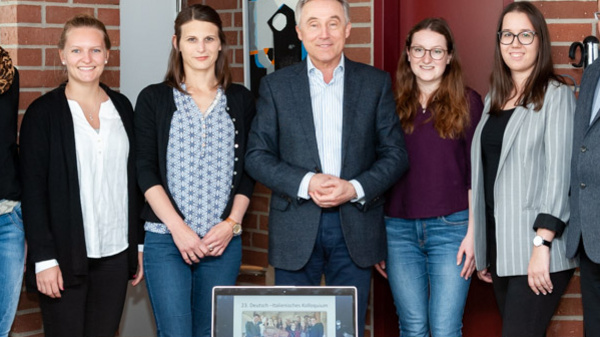 A group of six individuals stands in a bright office setting, with a laptop displaying a photo on a table in front of them. They are engaged in a collaborative atmosphere, dressed in casual professional attire, smiling, and positioned around a wooden table against a backdrop of brick walls and artwork.