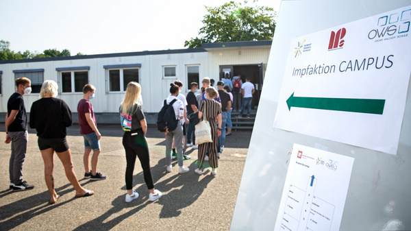 Un groupe de personnes attend dans une file pour entrer dans un bâtiment. Un panneau indique "Impfaktion CAMPUS" avec une flèche pointant vers la gauche, signalant l'emplacement d'une campagne de vaccination sur le campus.