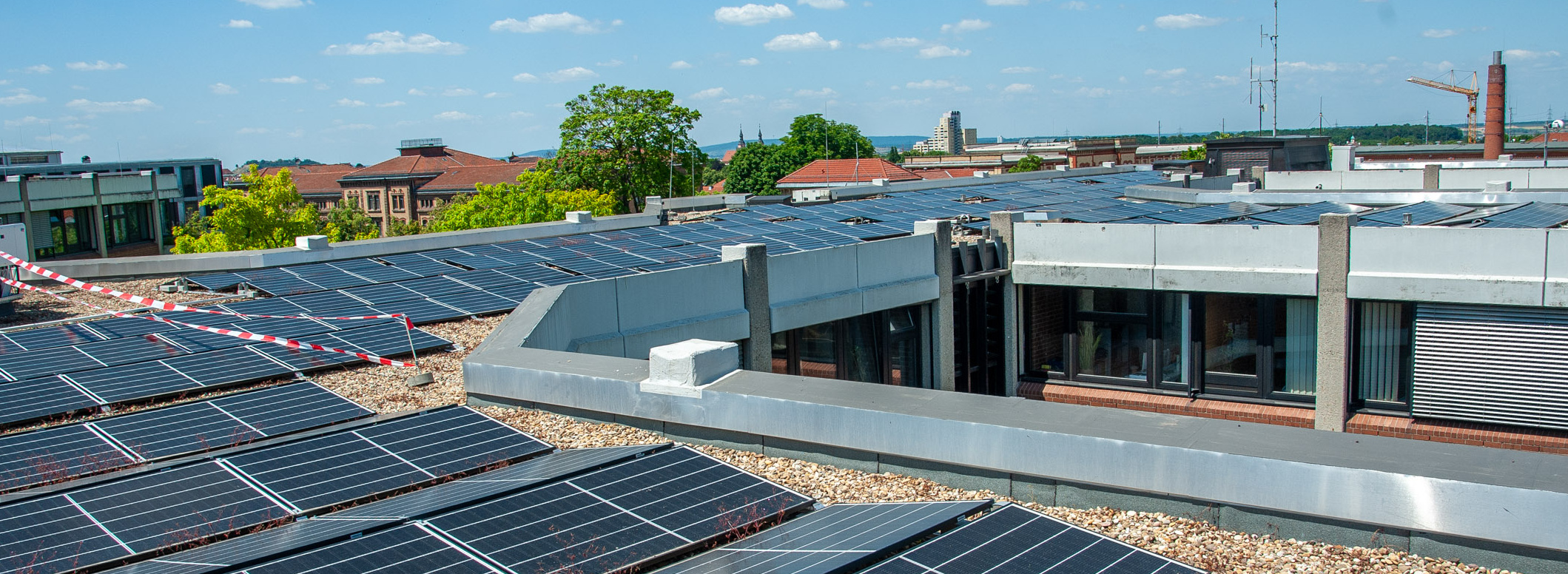 Header Bild: Eine Dachlandschaft mit mehreren Solarpaneelen, die in einem modernen städtischen Gebiet installiert sind. Helle Wolken am blauen Himmel schaffen eine sonnige Atmosphäre. Im Hintergrund sind Gebäude und Bäume sichtbar.