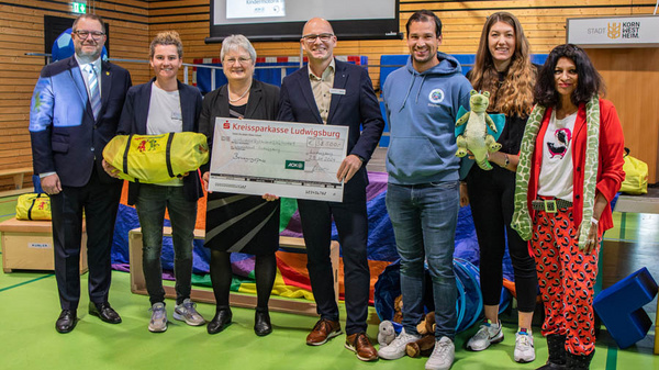 Eine Gruppe von sechs Personen steht in einer Sporthalle und hält einen großen Scheck. Die Personen lächeln und sind in Freizeitkleidung. Im Hintergrund sieht man Turnmatten und eine Basketballkorb. Der Scheck ist für die Kreissparkasse Ludwigsburg.