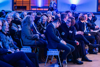 Eine Gruppe von Menschen sitzt bei einer Veranstaltung in einem gut beleuchteten Raum. Einige Personen interagieren miteinander, während andere auf ihre Plätze warten. Die Atmosphäre wirkt entspannt und gesellig. Die Stühle sind nummeriert, was auf eine organisierte Struktur hinweist.