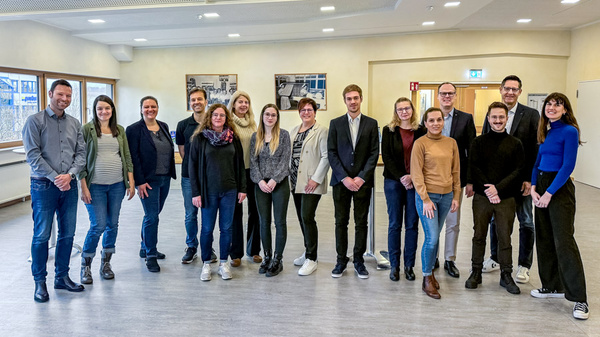 Eine Gruppe von 15 Personen steht in einem hellen Raum. Einige tragen formelle Kleidung, andere legere Outfits. Sie lächeln und posieren für das Foto. Im Hintergrund sind Bilder an den Wänden sichtbar. Der Raum wirkt freundlich und einladend.