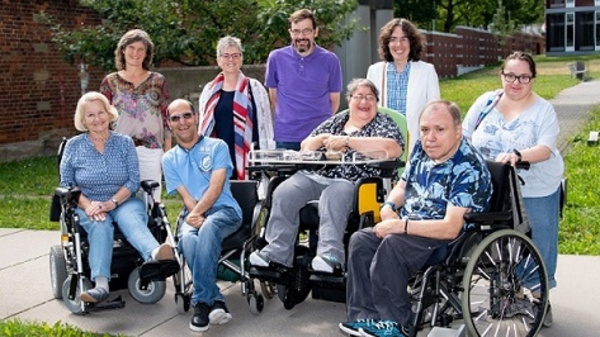 A diverse group of eight individuals, seated and standing, smiles for the camera. They use wheelchairs and vary in gender and age, showcasing an inclusive community. The background features greenery and a building, reflecting an outdoor setting.