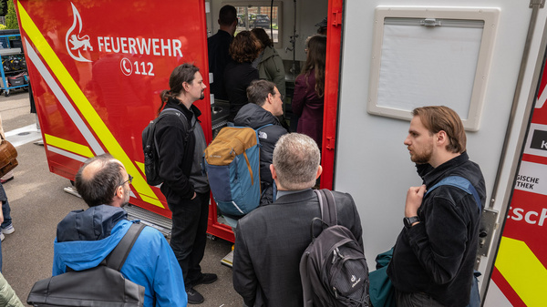 Eine Gruppe von Menschen steht vor einem Feuerwehrfahrzeug. Einige Personen sind im Gespräch, während andere in Richtung des Fahrzeugs schauen. Das Fahrzeug hat eine rote Außenseite mit dem Schriftzug "FEUERWEHR" und der Notrufnummer 112.