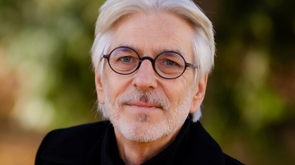 Portrait d'un homme d'âge moyen avec des cheveux mi-longs et argentés. Il porte des lunettes rondes et un manteau sombre. Son expression est douce, reflétant une attitude calme et accueillante, sur fond flou de verdure.
