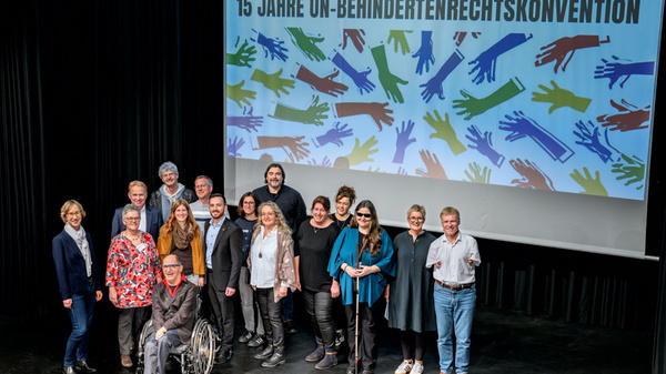 Die Aufnahme zeigt eine Gruppe von Menschen, die an einer Inklusionskonferenz teilnehmen, um das 15-jährige Bestehen der UN-Behindertenrechtskonvention zu feiern. Im Hintergrund befinden sich bunte Illustrationen von Händen, die Vielfalt und Inklusion symbolisieren.