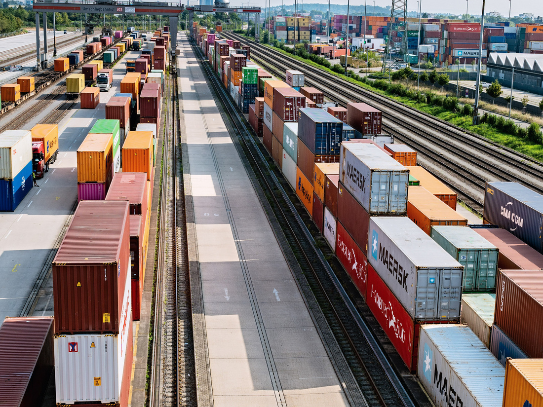 Ein Gleisbereich mit mehreren schiffbaren Containern in verschiedenen Farben. Die Container sind auf der linken und rechten Seite des Weges gestapelt, der in der Mitte verläuft. Die Szene zeigt eine aktive Lager- und Transportumgebung.