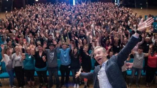 A large crowd of enthusiastic people is gathered in a hall, all raising their hands and smiling. In the foreground, a man with a joyful expression is posing for the photo, adding to the lively atmosphere of the event.