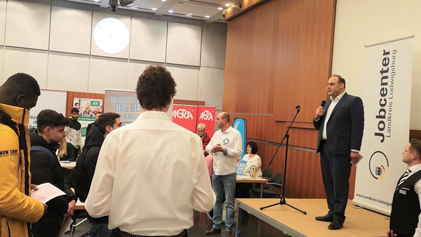 A speaker stands on a platform addressing an audience in a large room, with several attendees engaged and holding papers. Banners for job services are visible in the background, indicating a job fair or employment event.