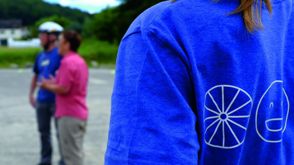 Un groupe de personnes se rassemble en pleine air pour une activité liée au vélo. Un individu, en t-shirt bleu, observe alors que deux autres personnes, l'une en rose et l'autre en vert, discutent avec un cycliste. L'environnement est calme et naturel.