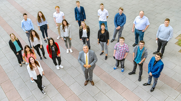 A diverse group of 20 individuals stands in a casual arrangement on a stone pavement. In the center, a man in a suit smiles, surrounded by men and women of various ages, dressed in a mix of casual and formal attire.
