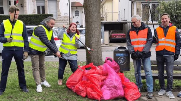 Gruppierung von sechs Personen in leuchtenden Warnwesten, die Müllsäcke halten. Sie stehen um einen Baum herum und wirken, als ob sie bei einer Aufräumaktion in einem Stadtpark mithelfen. Die Umgebung zeigt eine Wohngegend mit Gebäuden im Hintergrund.