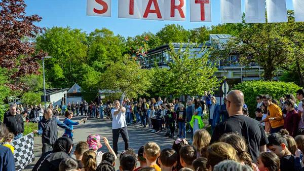 Eine Menschenmenge versammelt sich an einem Startpunkt eines Rennens, der mit einem großen, roten Schriftzug „START“ gekennzeichnet ist. Im Vordergrund stehen viele Kinder und Erwachsene, während einige aktiv für den Start des Rennens bereit sind. Bäume und Gebäude sind im Hintergrund sichtbar.