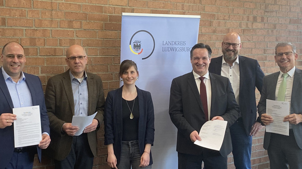 Sechs Personen stehen in einem Innenraum, einige halten Dokumente in der Hand. Im Hintergrund ist ein Banner mit dem Logo des Landkreises Ludwigsburg zu sehen. Die Personen lächeln und scheinen an einem offiziellen Anlass beteiligt zu sein, möglicherweise einer Unterzeichnung oder Bekanntgabe.
