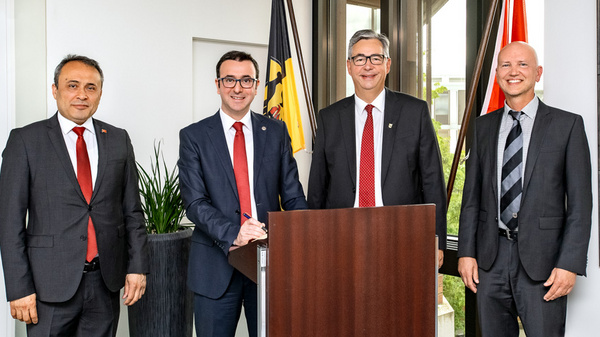 Four men in suits stand together, smiling. One man stands behind a podium, while the others are positioned beside him. They appear to be in an indoor setting with plants and flags visible in the background, suggesting a formal event or meeting.