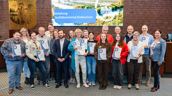 Eine Gruppe von Menschen steht in einem Raum, die meisten halten Urkunden in den Händen. Im Hintergrund ist ein Bildschirm mit dem Text „Verleihung Qualitätsmerkmal Kinderschutz“ zu sehen. Die Anwesenden wirken feierlich und stolz. (Text KI generiert)