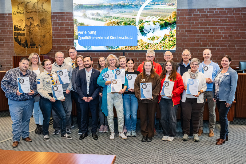 Eine Gruppe von Menschen steht in einem Raum, die meisten halten Urkunden in den Händen. Im Hintergrund ist ein Bildschirm mit dem Text „Verleihung Qualitätsmerkmal Kinderschutz“ zu sehen. Die Anwesenden wirken feierlich und stolz. (Text KI generiert)