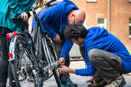 Deux hommes en sweatshirts bleus réparent un vélo. L'un est agenouillé près de la roue arrière, tandis que l'autre se penche pour l'aider. Une personne en arrière-plan regarde. L'environnement est urbain, avec des bâtiments en briques en arrière-plan. (Text KI generiert)