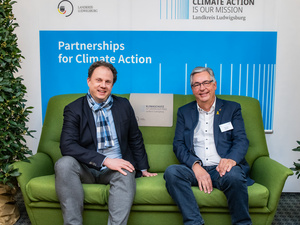 Zwei Männer sitzen auf einer grünen Couch und lächeln in die Kamera. Im Hintergrund steht ein Banner mit der Aufschrift „Climate Action is our Mission“ und „Partnerships for Climate Action“. Die Atmosphäre wirkt freundschaftlich und einladend.
