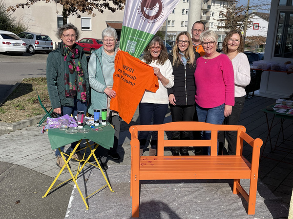 Un gruppo di otto persone sorride davanti a una panchina arancione. Alcuni di loro indossano magliette e le donne portano occhiali. Sul tavolo ci sono oggetti colorati. Un banner dietro di loro riporta un messaggio, mentre la scena si svolge all'aperto in un ambiente urbano.