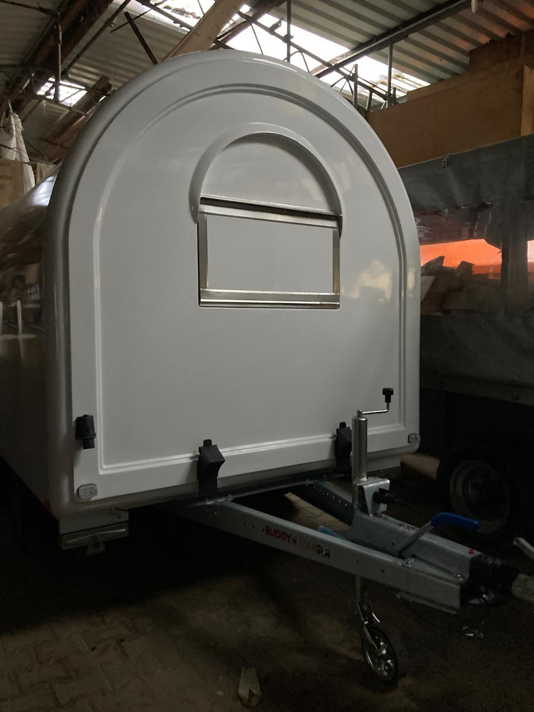 Ein weißer Anhänger mit einer gewölbten Rückseite und einem kleinen Fenster in der Mitte. Der Anhänger steht auf einem Auflieger und befindet sich in einer Halle. Es ist dunkel um den Anhänger herum, mit nur einem schwachen Lichtschein im Hintergrund.