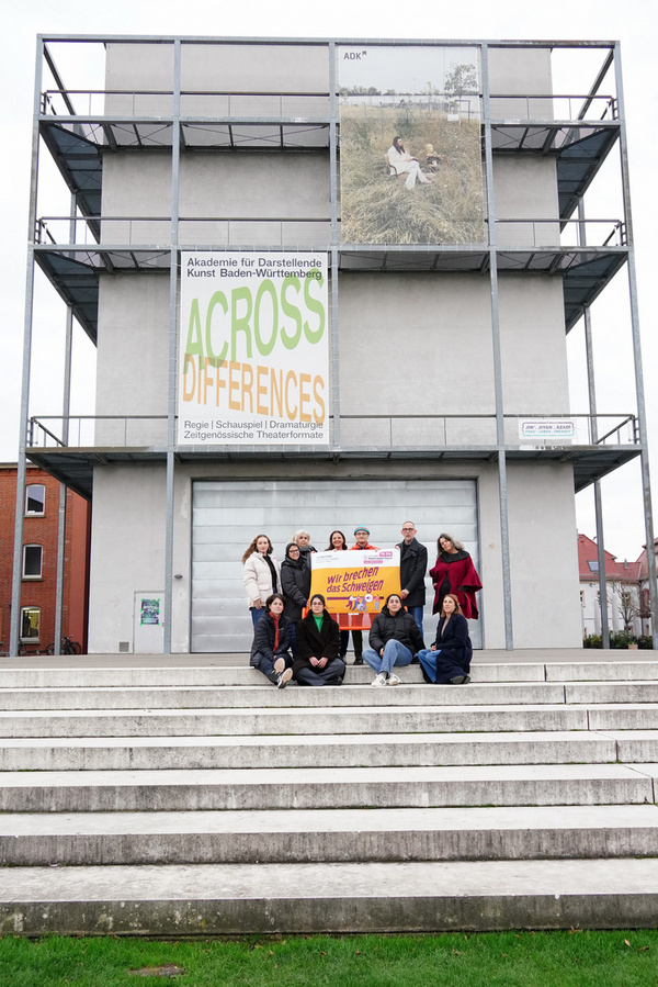 Un gruppo di persone è in piedi sui gradini di un edificio moderno, con una grande insegna che annuncia "ACROSS DIFFERENCES". Alcuni membri del gruppo tengono un cartello con un messaggio di unità. Lo sfondo mostra un cielo nuvoloso.