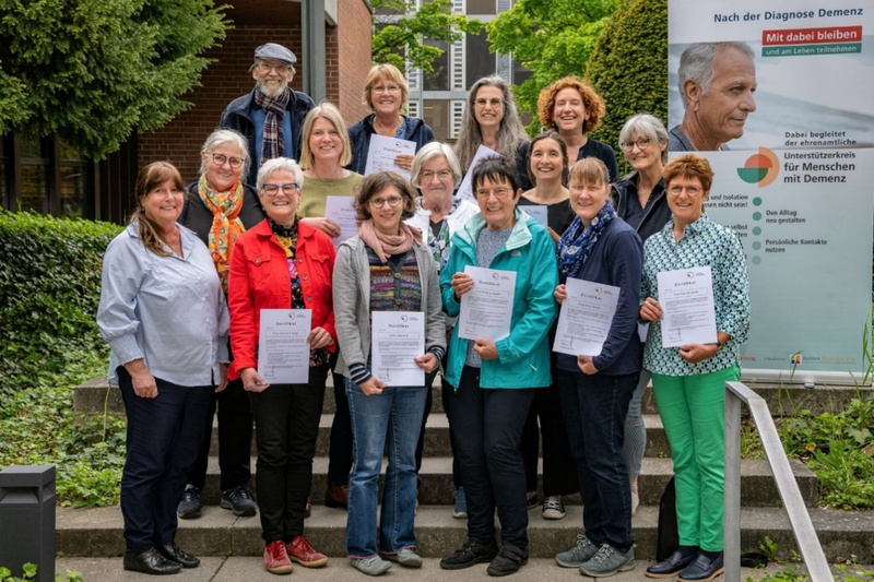 Eine Gruppe von 15 Personen steht im Freien und hält Urkunden in den Händen. Im Hintergrund befindet sich ein Banner mit Informationen zur Unterstützung von Menschen mit Demenz. Die Teilnehmerinnen und Teilnehmer lächeln und zeigen Stolz über ihre Leistungen.