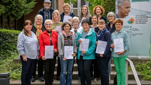Eine Gruppe von 15 Personen steht im Freien und hält Urkunden in den Händen. Im Hintergrund befindet sich ein Banner mit Informationen zur Unterstützung von Menschen mit Demenz. Die Teilnehmerinnen und Teilnehmer lächeln und zeigen Stolz über ihre Leistungen.