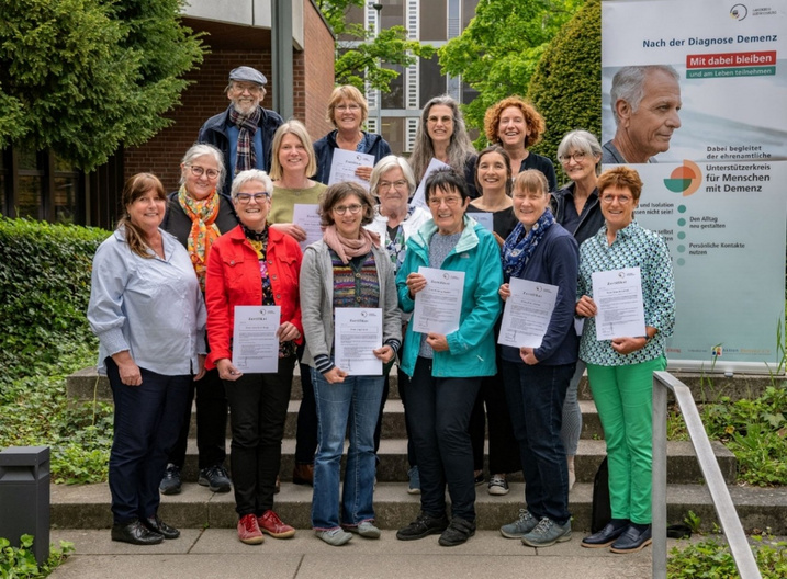 Eine Gruppe von 15 Personen steht im Freien und hält Urkunden in den Händen. Im Hintergrund befindet sich ein Banner mit Informationen zur Unterstützung von Menschen mit Demenz. Die Teilnehmerinnen und Teilnehmer lächeln und zeigen Stolz über ihre Leistungen.