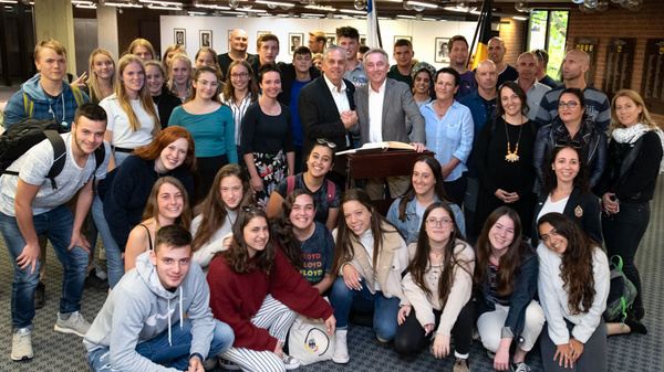A diverse group of around 40 people gather together in a hall, smiling and posing for a photo. They appear to be part of a community event or celebration, with some individuals in formal attire and others in casual clothing.