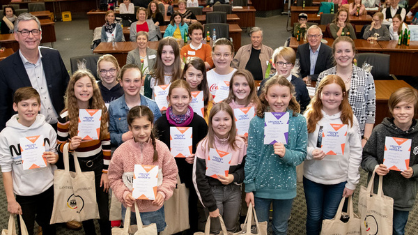 A group of enthusiastic children, holding awards and tote bags, stands in the foreground of a formal meeting room filled with audience members. The children are smiling and showcasing their accomplishments, while adults seated in the background observe the event.