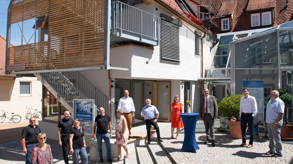 A group of twelve people stands outside a modern building with a wooden slatted upper section. The gathering includes men and women in diverse attire, with some seated and others standing. The setting features a cobblestone courtyard and blue sky overhead.
