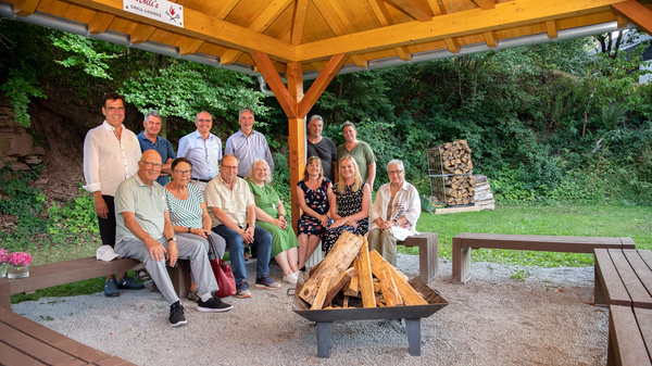 Eine Gruppe von 14 Personen sitzt in einem überdachten Bereich um ein Holzfeuer. Die Menschen lächeln und sind in legerer Kleidung. Im Hintergrund sind Bäume und ein Stapel mit Holzscheiten zu sehen. Es herrscht eine freundliche Atmosphäre.