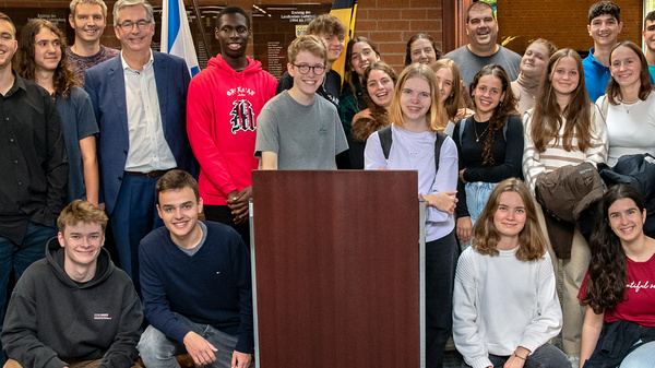 Groupe d’étudiants réunis autour d’un pupitre dans une salle. Ils sourient et posent ensemble, montrant une bonne ambiance. Quelques adultes sont également présents, indiquant un cadre formel ou événement académique. L'image reflète l'unité et la collaboration.