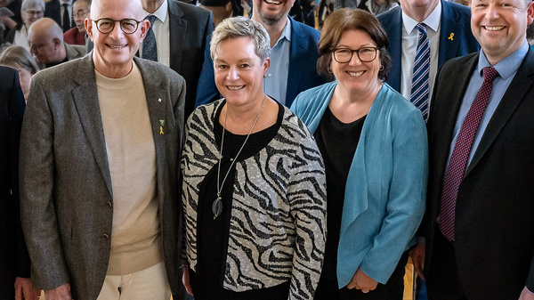 Eine Gruppe von acht Personen steht lächelnd vor einem Publikum in einem Saal. Die Personen sind in formeller Kleidung gekleidet. Im Hintergrund sind viele Menschen zu sehen, die auf Stühlen sitzen. Die Atmosphäre wirkt freundlich und einladend.