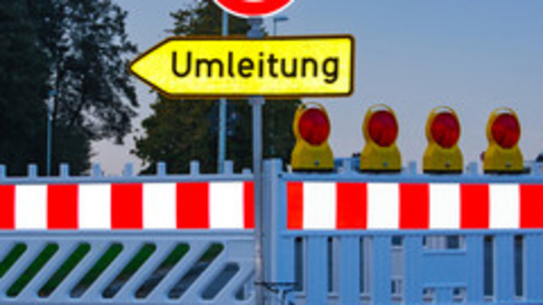 A roadblock is featured with a red and white barrier, and a circular road sign indicating 'no entry.' There is a yellow sign pointing to the right labeled "Umleitung," which means "detour" in German. Several warning lights are positioned at the top of the barrier.